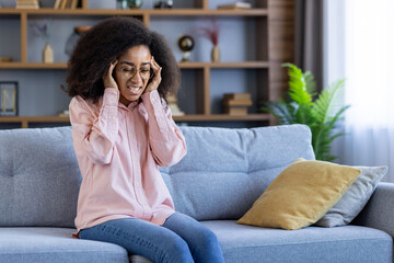 A woman sitting on a couch at home, holding her head in pain and discomfort. She appears distressed while being in a domestic setting.