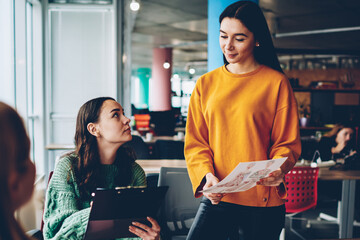 Young women talking about new design sketch during brainstorming in office interior.Skilled students collaborating on blueprint of fashion clothes sitting at desktop in stylish studio