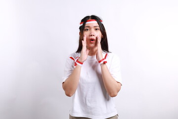 Young Asian woman wearing red white flag ribbon shouting loud at the camera, telling about good sale offer isolated over white background. Celebrate Indonesian independence day on 17 August