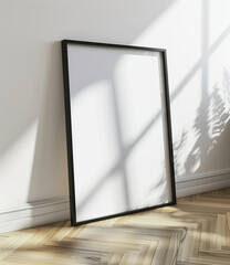 Empty black frame leaning against a white wall in a sunlit room with herringbone wooden floor. Minimalist interior mockup for design and print.
