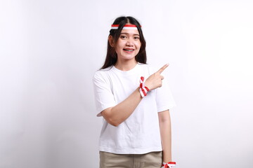 Happy young Asian woman in red white flag ribbon pointing fingers aside at copy space isolated on white background. Indonesian independence day on 17 august concept