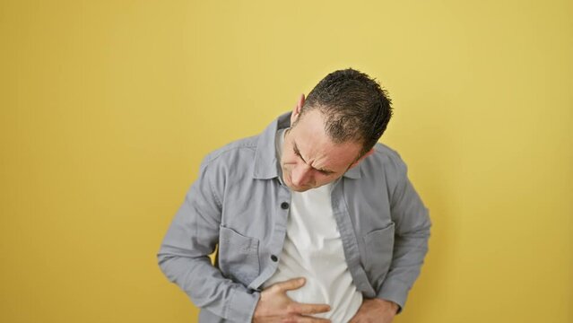 Attractive young hispanic man in pain, clutching stomach with a grimace. nausea from a cruel bout of illness, standing isolated over a yellow background