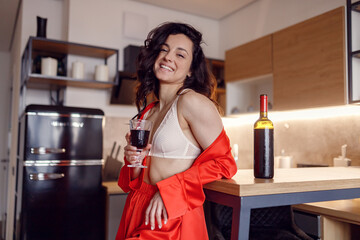 Smiling sexy lady wearing silky red pajamas while holding glass of wine, posing at the kitchen at home, looking at camera. Rest time concept