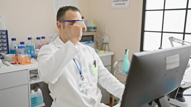 A focused hispanic man wearing safety goggles works at a computer in a modern laboratory setting, representing professionalism in healthcare.