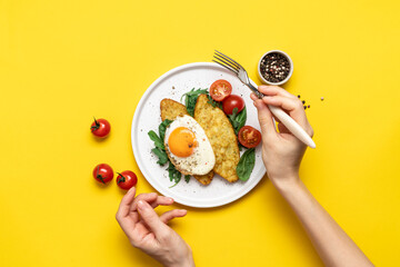 Female hands with fork on the background of plate with breakfast on bright yellow background. Egg,...