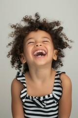 young mixed-race child with curly hair, wearing a black and white striped dress with ruffled straps, smiling and laughing in a minimalist studio setting with a plain white background