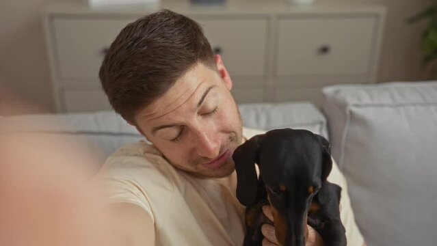 A young hispanic man sits in his living room holding a dachshund, capturing a selfie in a comfortably furnished apartment