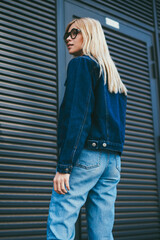 Rear view of stylish young woman dressed in denim clothes standing on black promotional background...