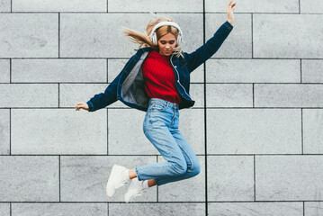 Stylish hipster girl dressed in trendy outfit jumping while listening cool electronic music in...