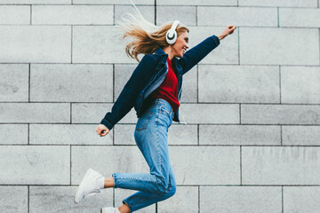 Cheerful young woman dressed in casual denim apparel jumping outdoors on grey wall.Positive hipster...