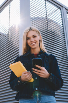 Half length portrait of pretty smiling blonde blogger sending funny messages to friends via modern smartphone.Gorgeous happy teenager looking at camera while installing application on telephone