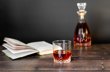 There is a decanter and a glass of cognac on the wooden table and there is an open book