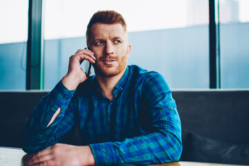 Handsome young man with red hair talking with friends on smartphone device resting in coworking space.Hipster guy dressed in casual shirt communicating on mobile phone sitting in coffee shop