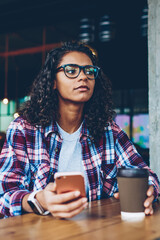 African american hipster girl in eyewear resting with coffee to go during break while sending sms...