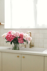 Beautiful peonies in vase in sink on background of brass faucet and window in new scandinavian house. Pink peony flowers in modern kitchen interior, summer floral arrangement