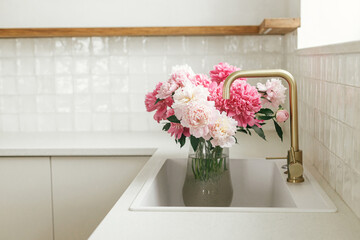 Beautiful peonies in vase in sink on background of brass faucet and window in new scandinavian...