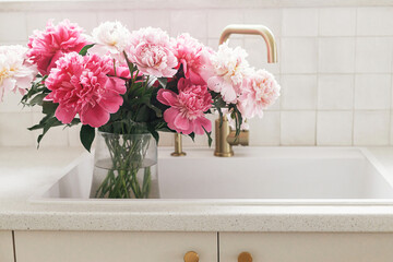 Beautiful peonies in vase in sink on background of brass faucet and white counter in new scandinavian house. Pink peony flowers in modern kitchen interior, summer floral arrangement