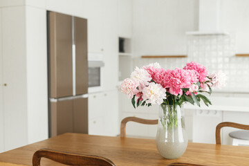 Beautiful peonies in vase on wooden table on background of stylish white kitchen with appliances in new scandinavian house. Modern kitchen interior and summer floral arrangement