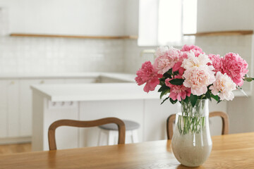 Beautiful peonies in vase on wooden table on background of stylish white kitchen with island, wooden shelves and appliances in new house. Modern kitchen interior in farmhouse