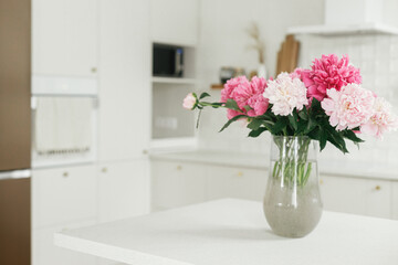 Beautiful peonies in vase on granite countertop on background of stylish white kitchen with wooden shelves and appliances in new house. Modern minimal kitchen interior