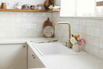Modern white kitchen with brass faucet, granite countertop, wooden shelves with utensils and roses in new scandinavian house. Modern kitchen interior. Modern minimal kitchen design.