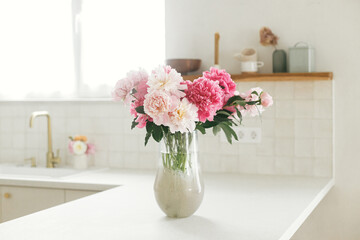 Beautiful peonies in vase on granite countertop on background of stylish white kitchen with wooden shelves and appliances in new house. Modern minimal kitchen interior