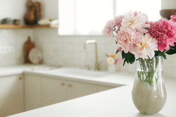 Beautiful peonies in vase on granite countertop on background of stylish white kitchen with wooden shelves and appliances in new house. Modern minimal kitchen interior