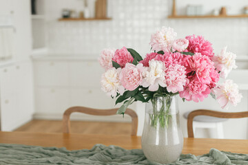 Beautiful peonies in vase on wooden table on background of stylish white kitchen with appliances in new scandinavian house. Minimal kitchen design. Modern kitchen interior