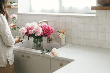 Woman arranging beautiful peonies in vase at sink with brass faucet and granite countertop. Florist...