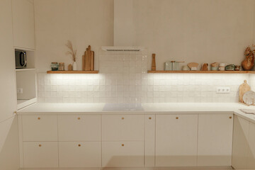 Modern kitchen interior. Stylish white kitchen cabinets with brass knobs, wooden shelves with evening light and appliances in new scandinavian house. Modern minimal kitchen design