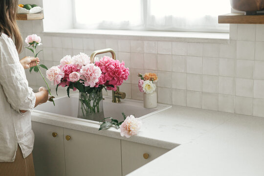 Woman arranging beautiful peonies in vase at sink with brass faucet and granite countertop. Florist holding pink peony and scissors in new modern kitchen, hands with flowers close up