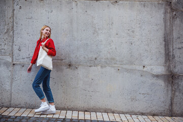 Young cheerful blonde girl in sunglasses and red jacket posing near a gray wall