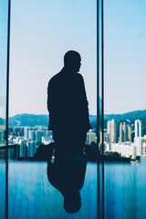 Fototapeta premium Back silhouette of owner of company standing near big glass windows and looking at amazing evening scenery of modern city with skyscrapers and high mountaons on background after hard working day