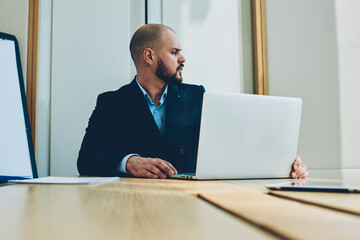 Confident male trader looking away while working at modern laptop computer connected to wireless...