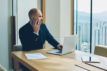 Confident proud ceo in formal clothes talking with manager about accounting reports on smartphone working on developing business project at laptop computer connected to wireless internet in office