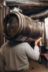 Young man in leather apron holding beer keg at modern brewery, craft brewery worker