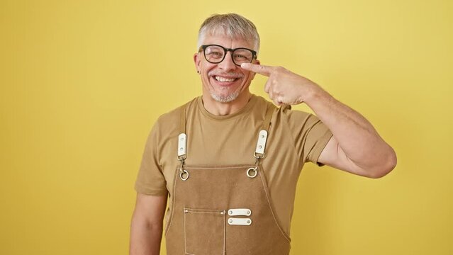 Cheerful middle age grey-haired man, wearing glasses and apron, ecstatically pointing at his fresh face and nose, promoting beauty health on a bright yellow isolated background