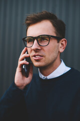 Close up image of young handsome man in cool spectacles communicating with friends via application on smartphone connected to 4G.Good looking hipster guy talking on cellphone during walk at street