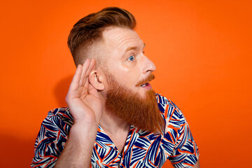 Photo of curious funky guy dressed print shirt arm ear listening secret isolated orange color...