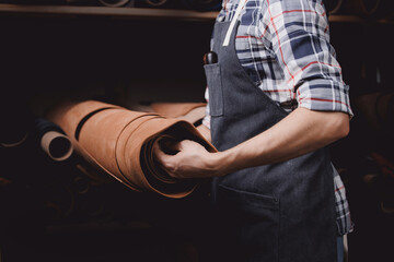 Cobbler Man tailor chooses roll of brown leather for sewing a bag or shoes made of skin in workshop