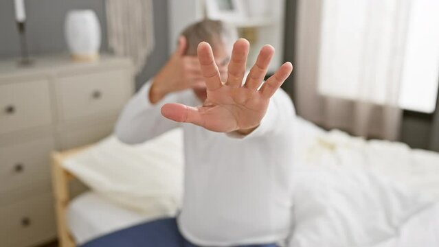 Embarrassed middle-aged grey-haired man in pyjamas fearfully covers eyes with hands, gesturing stop in bedroom