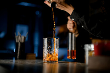 Bartender pours a brown alcoholic drink from a jigger into a tall cocktail glass