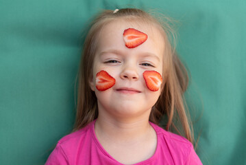 A young girl with strawberry slices on her face smiles while sitting on a teal background