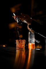 Bartender's hands pour from a jigger the last drops of alcoholic drink into a low glass