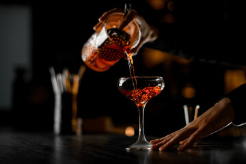 Hands of a bartender pouring a cocktail from a mixing glass, straining it through a sieve into a...