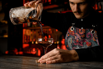 Bartender bends over and pours a brown cocktail from a mixing glass into a tall stemmed glass