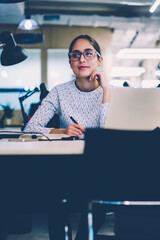 Pensive female in eyewear pondering on creating business plan for startup using laptop computer for...