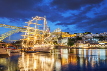 Fototapeta premium View of Vila Nova de Gaia city with a sailing ship and Dom Luis I bridge over Douro river in the evening. Porto, Portugal