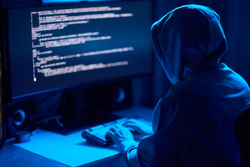 In a dimly lit room, a cloaked figure types code on a computer, hinting at a mysterious hacker