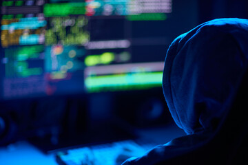 In a dimly lit room, a cloaked figure types code on a computer, hinting at a mysterious hacker